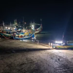 Sembilan unit kapal nelayan asal Muncar sedang bersandar dan melakukan perbaikan di pinggir pantai Resor Sembulungan pada malam hari dengan pencahayaan lampu kapal.