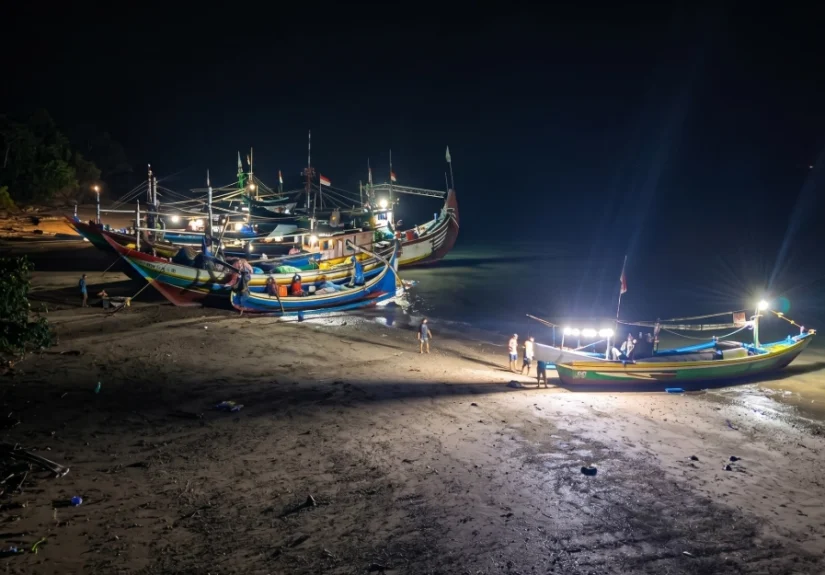 Sembilan unit kapal nelayan asal Muncar sedang bersandar dan melakukan perbaikan di pinggir pantai Resor Sembulungan pada malam hari dengan pencahayaan lampu kapal.